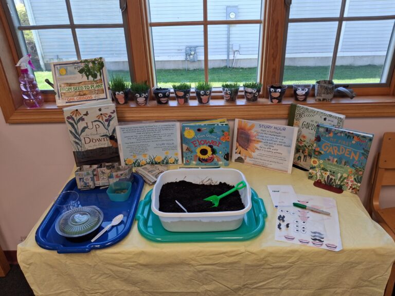 A library table is set up with a children’s gardening activity, including a bin of soil, small containers, a spoon, and planting supplies. Around the table are picture books about plants, a sign with sunflower‑planting instructions, and decorated grass‑head cups growing on the windowsill behind the display.