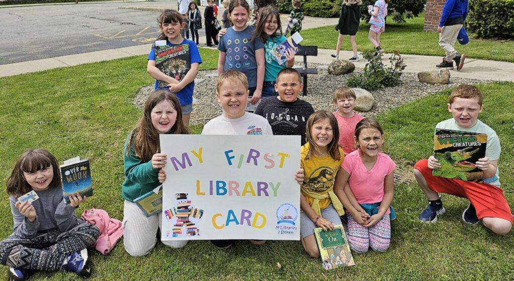 A group of children sit and stand on a grassy area outside, smiling and holding books and a large colorful sign that reads “My First Library Card.” Behind them are a sidewalk, landscaped rocks, and other children walking nearby, creating a cheerful scene celebrating reading and library participation.