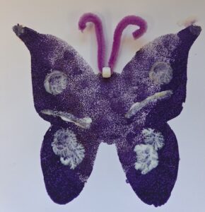 A purple butterfly-shaped craft made from textured foam or sponge is shown on a plain white background. The wings have irregular white painted patches, and two pink pipe cleaners with a small white bead between them form the antennae and head.