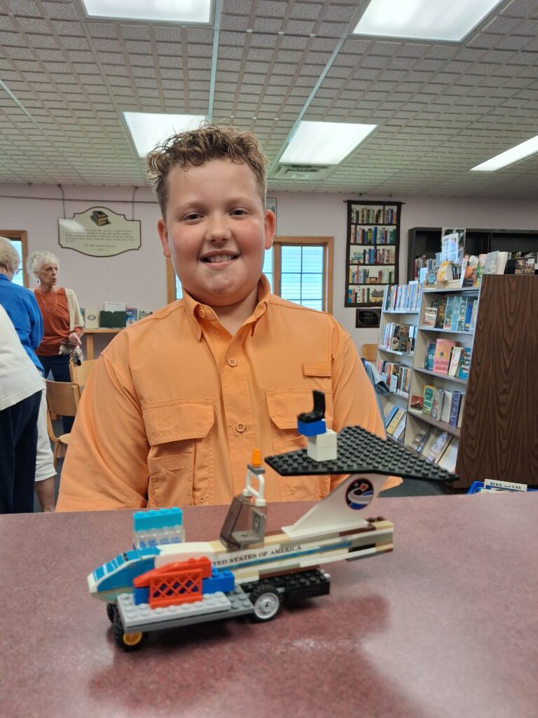 The image shows Caleb, a smiling 9-year-old boy wearing an orange button-up shirt seated at a table inside a library. In front of him is a detailed LEGO model of a space shuttle labeled “United States of America,” built with white, blue, orange, and black pieces, while bookshelves and other patrons appear in the background.