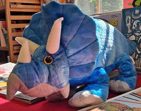 A plush toy shaped like a triceratops dinosaur sits on a red table surrounded by books. The toy is bright blue with lighter blue and gray accents, three white horns, and shiny golden eyes, creating a cheerful and inviting display in a well-lit library or reading area.