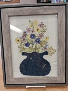 The image shows a framed piece of textile art featuring a dark blue appliquéd vase with embroidered flowers and leaves extending outward. The flowers are stitched in shades of yellow, purple, and pink with green leaves, displayed under glass in a light brown wooden frame on a tabletop.