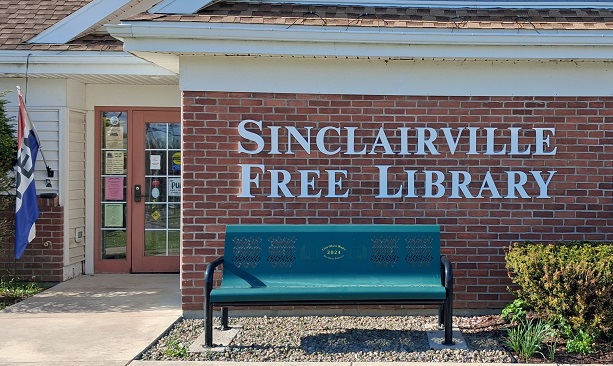 The image shows the front entrance of the Sinclairville Free Library building. The brick exterior features large white lettering spelling the library’s name above a green metal bench and a glass door with posted notices and a flag nearby, creating a welcoming community space.