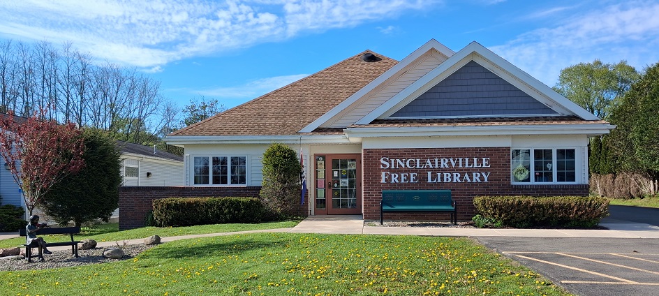 The image shows the Sinclairville Free Library building on a clear, sunny day. The one-story structure has a brown shingled roof, white trim, and a brick front with large white lettering spelling the library’s name; a green bench sits outside near the entrance, surrounded by neatly trimmed bushes and a small garden with a sculpture of a person reading.