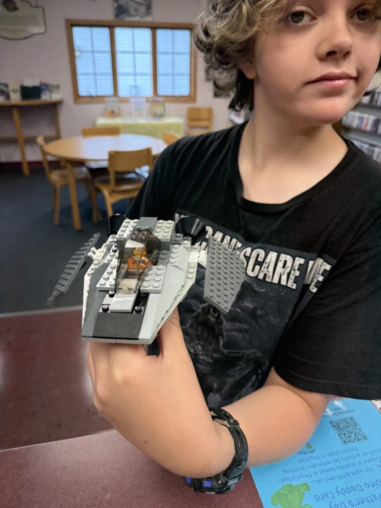 The image shows James, a 12-year-old boy with curly hair wearing a black T-shirt and wristwatch, holding a gray LEGO spacecraft model. The detailed build features angled wings, a cockpit with a small LEGO figure inside, and is displayed in a library setting with tables, chairs, and shelves visible in the background.