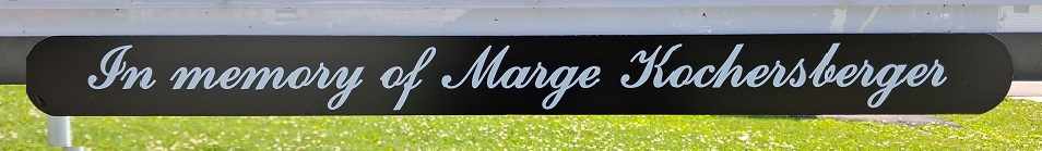 The image shows a black rectangular plaque with rounded edges mounted outdoors. It features white cursive lettering that reads “In memory of Marge Kochersberger”, set against a background of green grass, indicating a memorial dedication attached to a public sign.