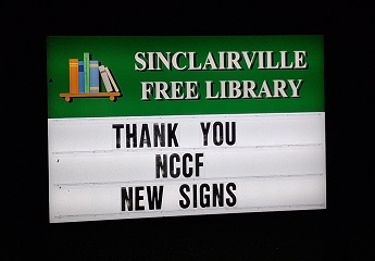 The image shows an illuminated outdoor sign for the Sinclairville Free Library at night. The green header features colorful books and white text with the library’s name, while the message board below reads “THANK YOU NCCF NEW SIGNS,” expressing gratitude to the Northern Chautauqua Community Foundation for supporting new signage.