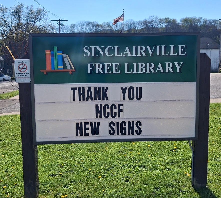 The image shows an outdoor sign for the Sinclairville Free Library standing on a grassy area with dandelions. The sign has a green header with colorful books and white text reading “Sinclairville Free Library,” and the message board below says “THANK YOU NCCF NEW SIGNS,” expressing appreciation to the Northern Chautauqua Community Foundation for the library’s new signage.