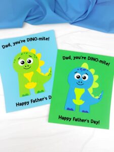 Two colorful Father’s Day cards feature cheerful cartoon dinosaurs made from cut-out paper shapes. One card shows a green dinosaur on a light blue background, and the other a blue dinosaur on a green background, both with the message “Dad, you’re DINO-mite! Happy Father’s Day!” in black text.