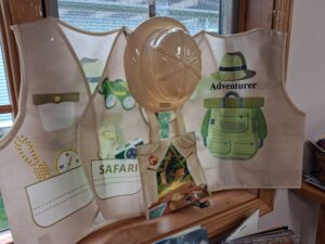 The image shows a display near a window featuring two light-colored fabric vests and a beige safari-style helmet. One vest is labeled “Safari” with illustrations of a compass, rope, binoculars, and camera, while the other reads “Adventurer” with a green backpack and hat, creating a playful exploration-themed setup for a children’s program or activity.