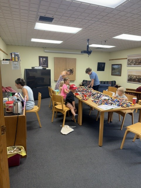 Several children and adults gather around long tables covered with colorful LEGO bricks in a library meeting room. The space has beige walls, framed photos, and wooden chairs, showing a lively creative building session.