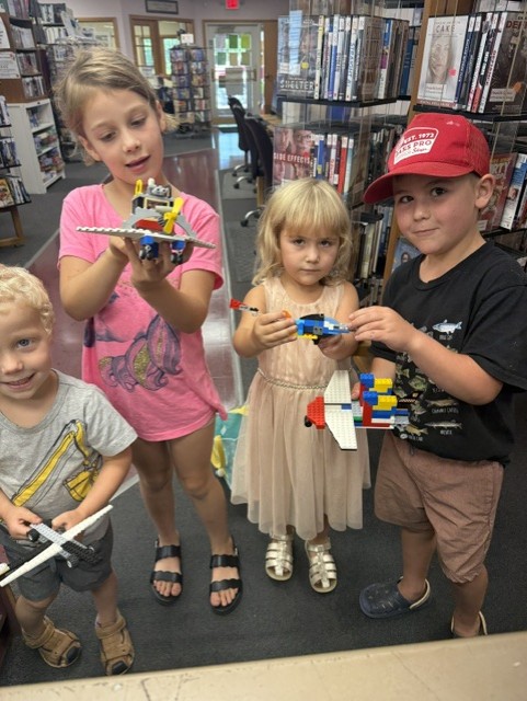 Four young children stand in a library holding colorful LEGO models shaped like airplanes and vehicles. Bookshelves and a doorway are visible behind them, and the children smile proudly at their finished projects.