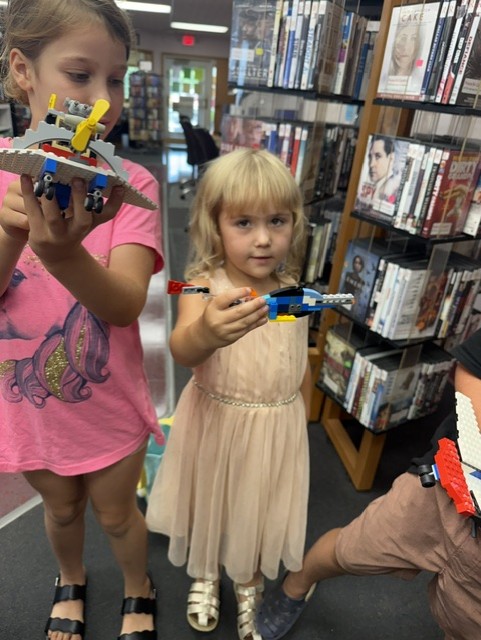 A young girl in a light pink dress holds a small blue and red LEGO model while standing beside another child in a pink shirt. They are in a library aisle surrounded by shelves of DVDs and books.