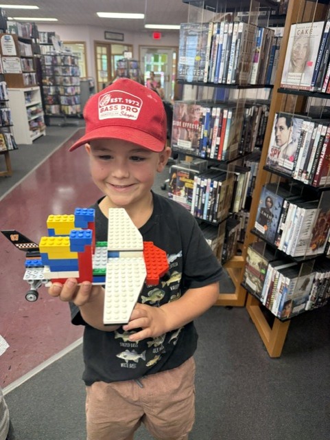 A boy wearing a red Bass Pro Shops hat and black shirt smiles while holding a colorful LEGO airplane. He stands in a library with shelves of DVDs and books in the background.
