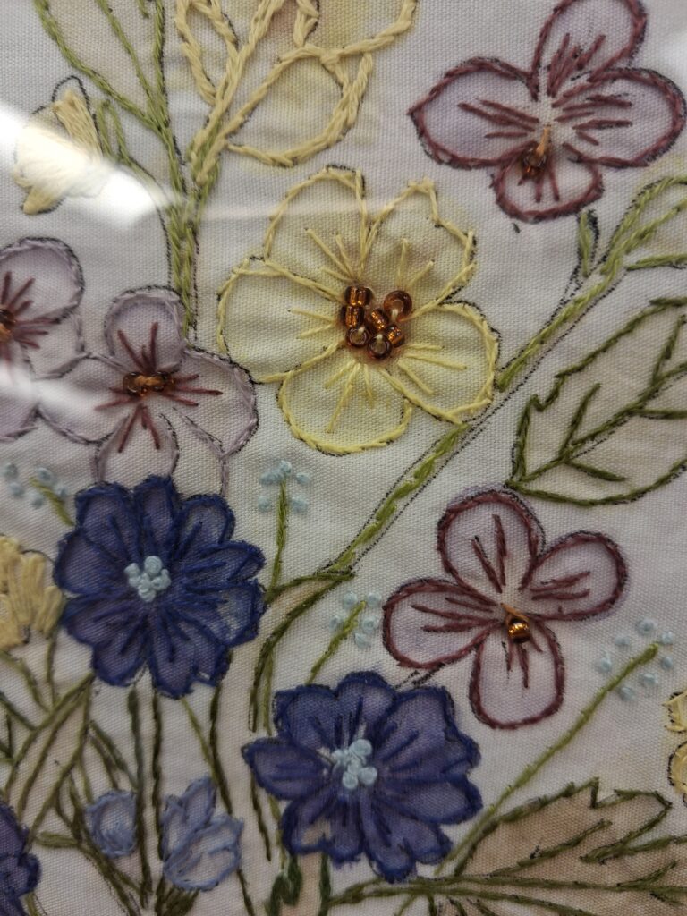 The image shows a close-up of embroidered fabric with a floral design featuring yellow, purple, and blue flowers stitched with green stems and leaves. Some flower centers are decorated with small golden beads, highlighting the detailed threadwork and texture against a light background.