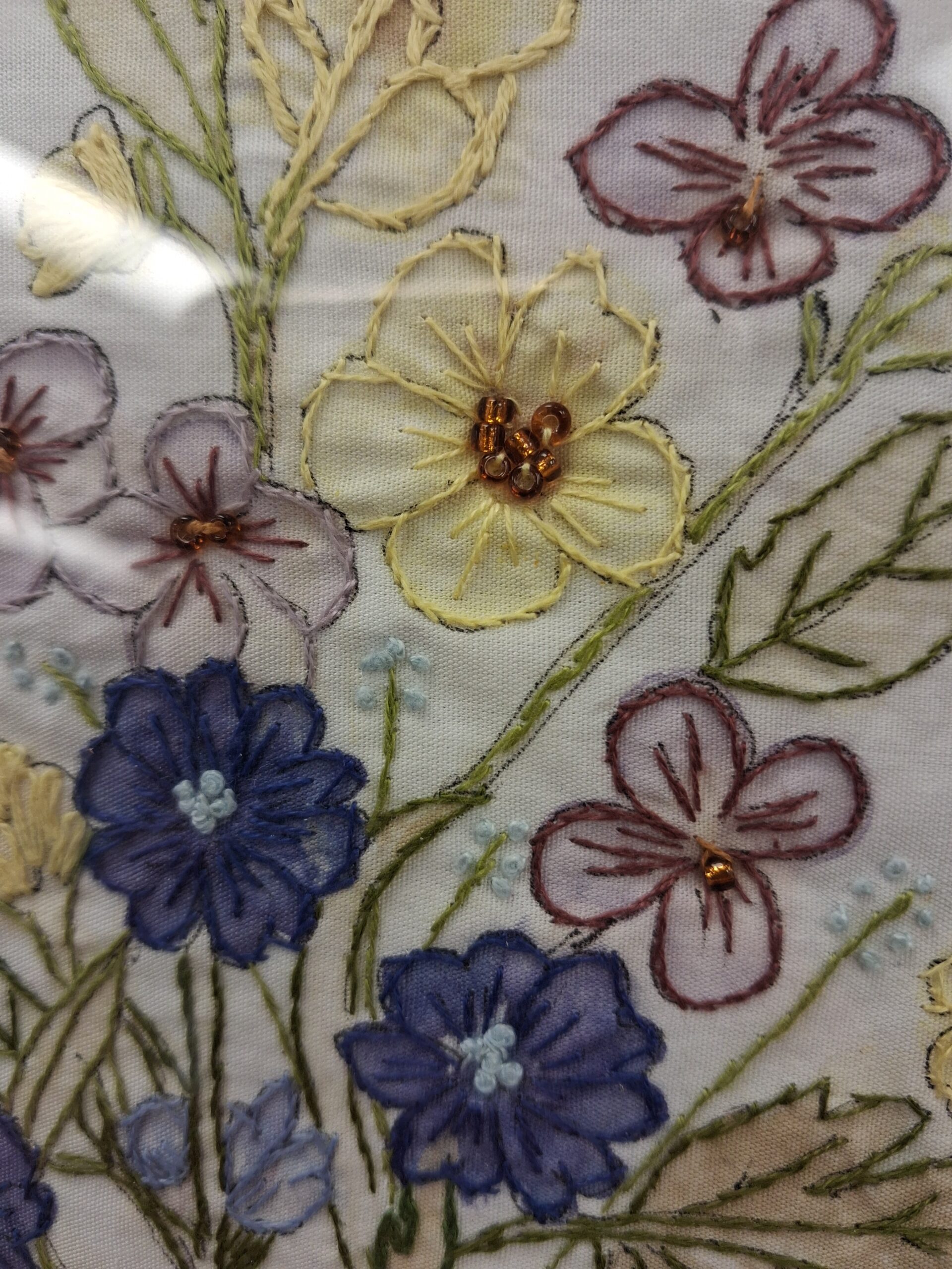 The image shows a close-up of embroidered fabric with a floral design featuring yellow, purple, and blue flowers stitched with green stems and leaves. Some flower centers are decorated with small golden beads, highlighting the detailed threadwork and texture against a light background.