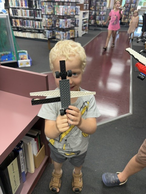 A small boy with curly blond hair holds a simple black and white LEGO airplane in front of his face. He stands near a pink shelf in the library, with other children visible in the background.