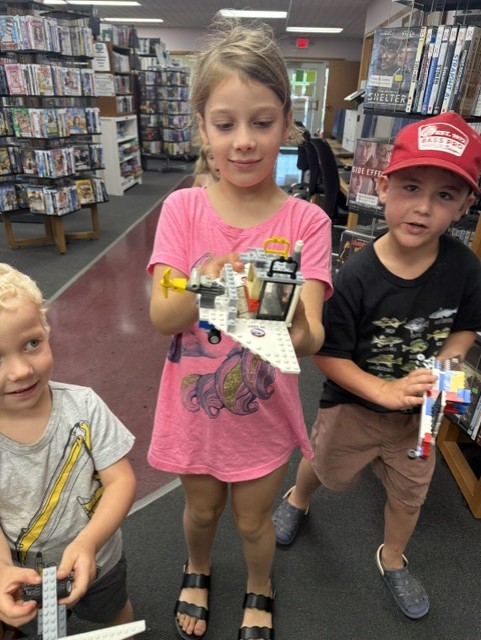 Three children stand together in a library aisle, each holding a unique LEGO creation made from multicolored bricks. They smile and show off their imaginative designs amid shelves of DVDs and books.