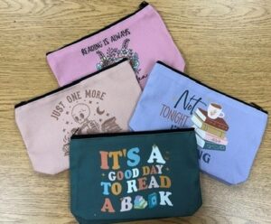 The image shows four colorful fabric pouches arranged on a light wood table. Each pouch features a different book-themed design and phrase, including “It’s a good day to read a book” on a dark green pouch, “Not tonight” with a stack of books on a lavender pouch, “Just one more” on a light pink pouch, and “Reading is always” on a mauve pouch, celebrating reading and library culture.